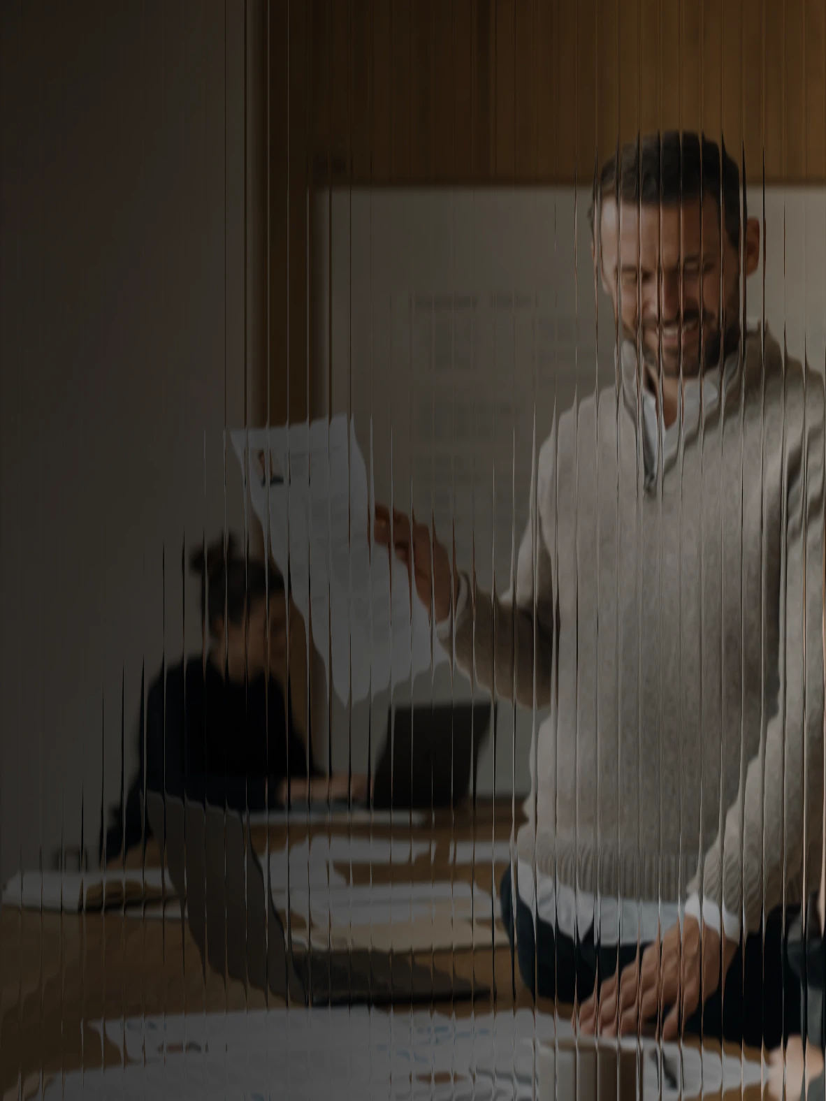 A recruiter smiling as he holds up a CV for a colleague, past applications spread along the table, seen through fluted glass